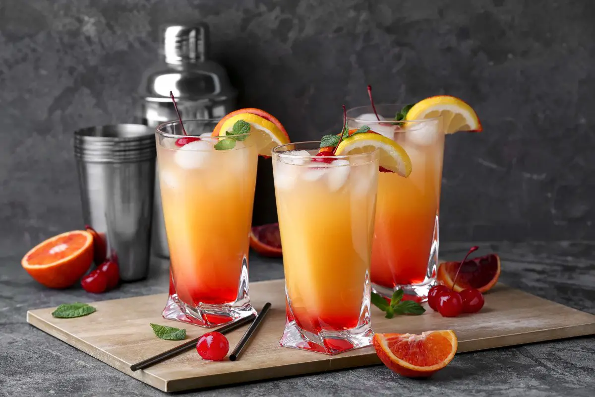 Three tequila sunrise cocktails with cherry orange garnish and cocktail shaker